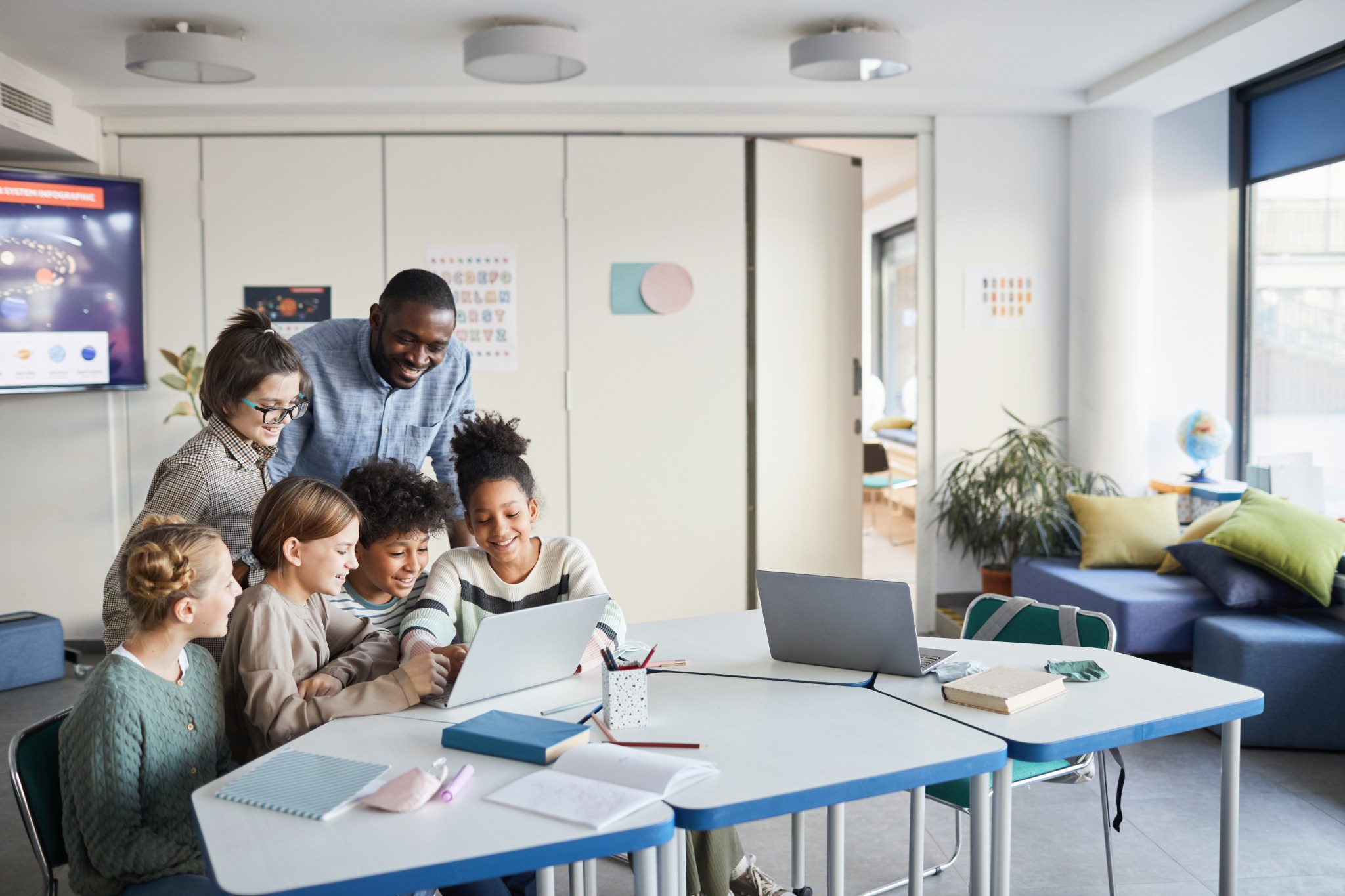 A teacher and a diverse group of students gather around a laptop in a modern classroom.